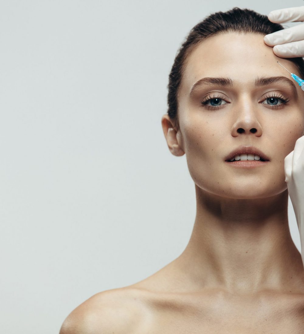Woman having injection on forehead. Anti wrinkles aesthetic surgery in forehead done by cosmetic doctor on grey background.